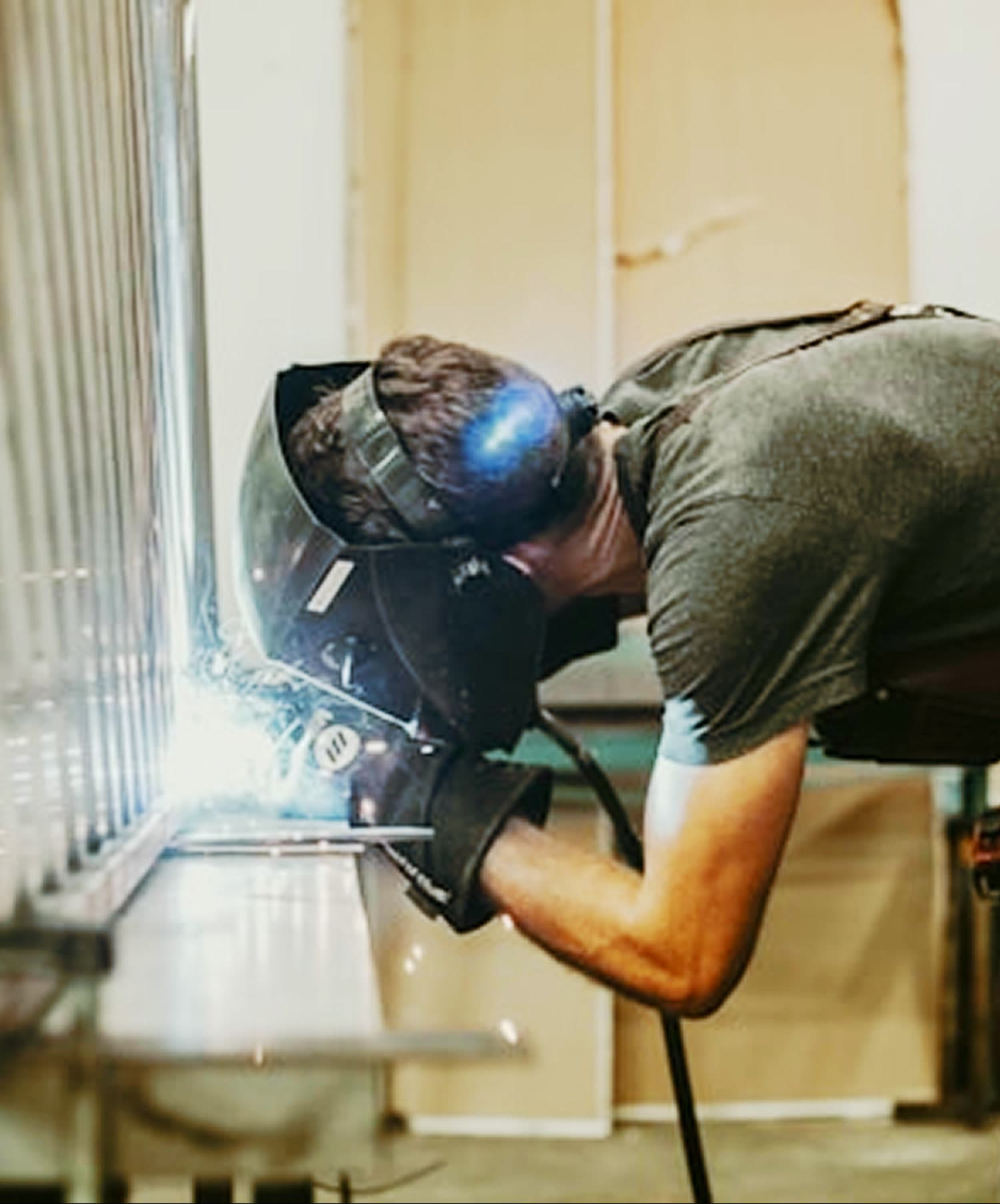 A welder bent over a bench in a helmet, joining a metal frame as sparks scatter.